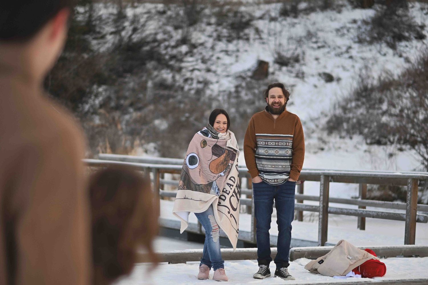Couple wrapped in a decorative blanket standing together on a snowy bridge during a winter outdoor photoshoot.