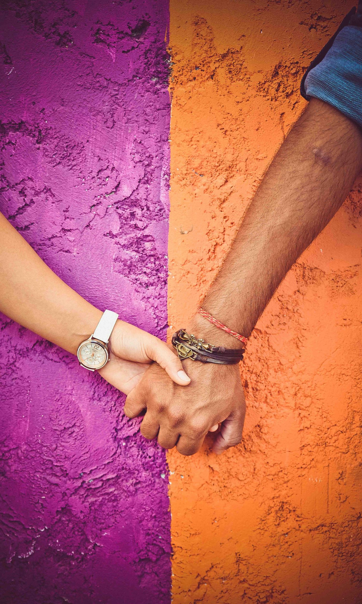 A couple holds hands against a vibrant split purple and orange textured wall, wearing a watch and leather bracelet.