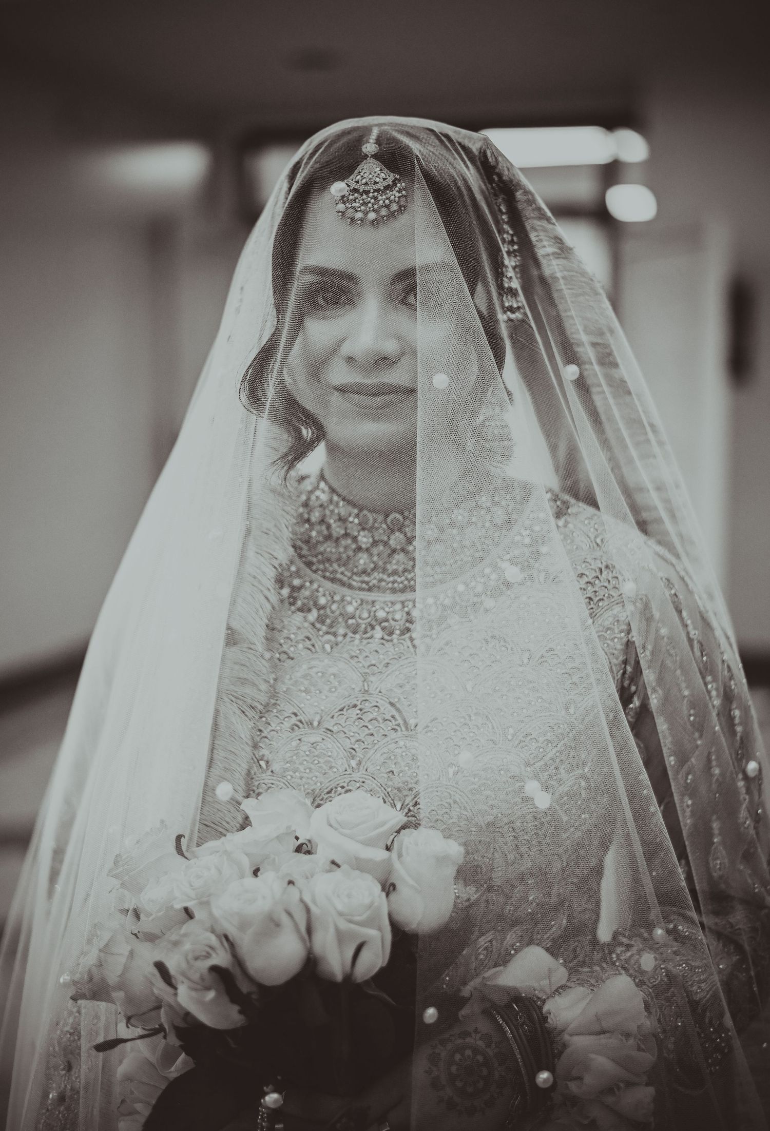 Black and white portrait of a South Asian bride in traditional attire, wearing a veil and holding white roses.