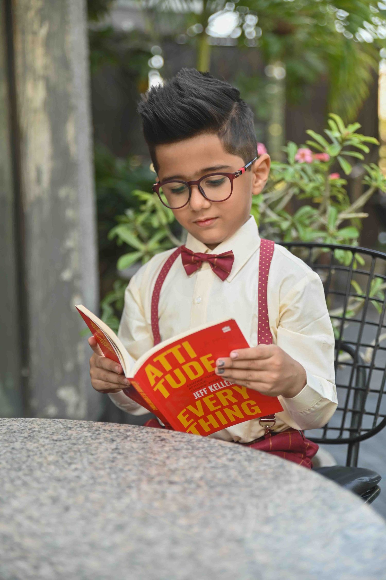 Stylish boy in glasses, bow tie and suspenders reading Attitude is Everything book outdoors.