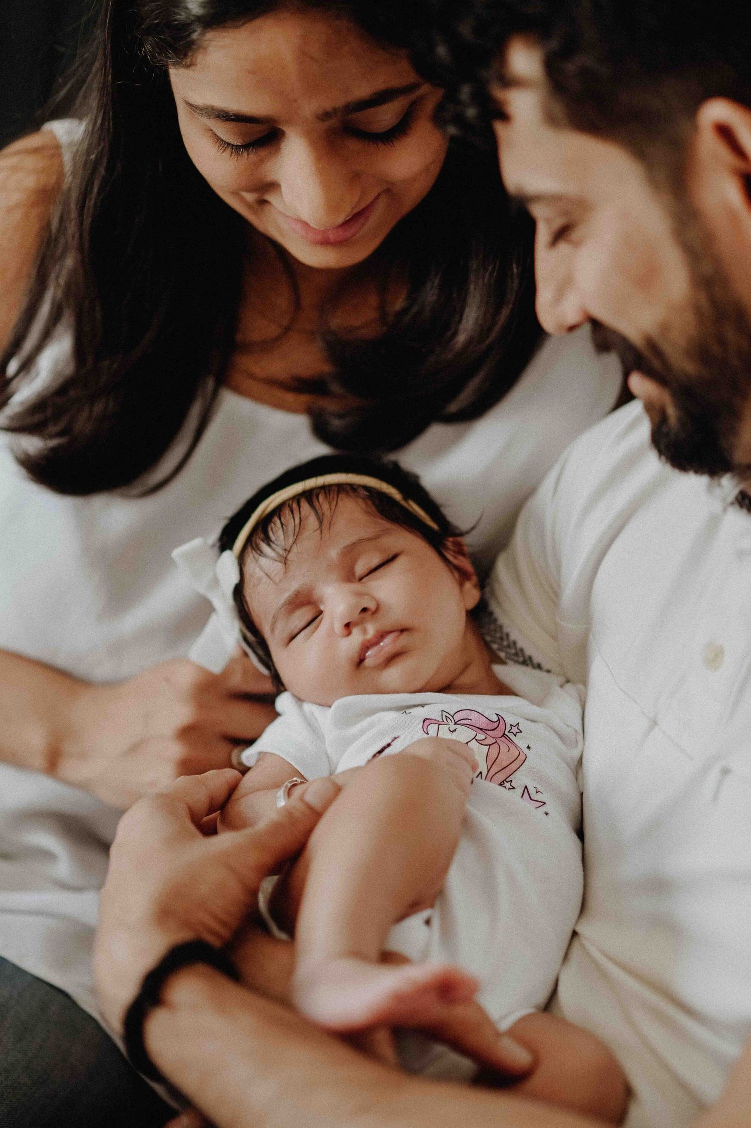New parents smile lovingly at their sleeping newborn baby in white outfits.