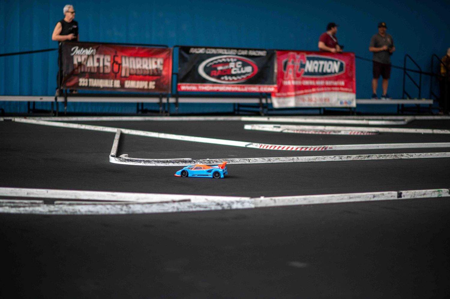 Blue and orange RC car racing on an indoor black track with sponsor banners.