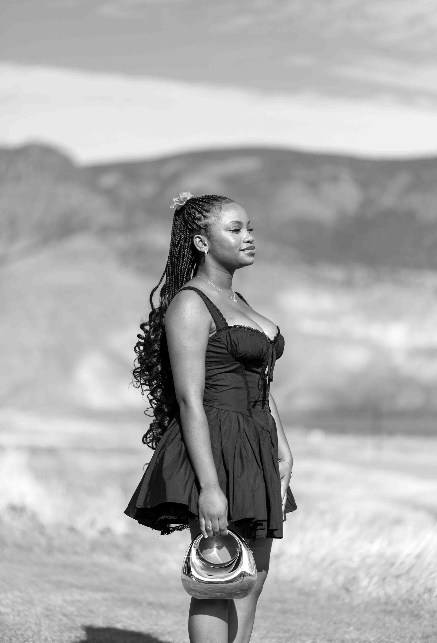 Black and white portrait of woman in black dress holding metallic bag with mountain scenery.