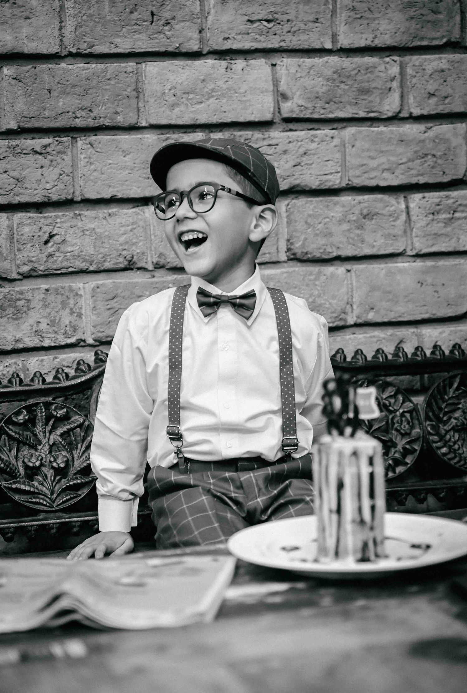 A joyful young boy in retro suspenders, bow tie, and newsboy cap laughs beside a birthday cake.