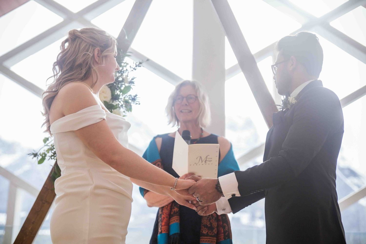 Bride and groom exchange vows holding hands before officiant inside bright modern wedding venue with mountain views.