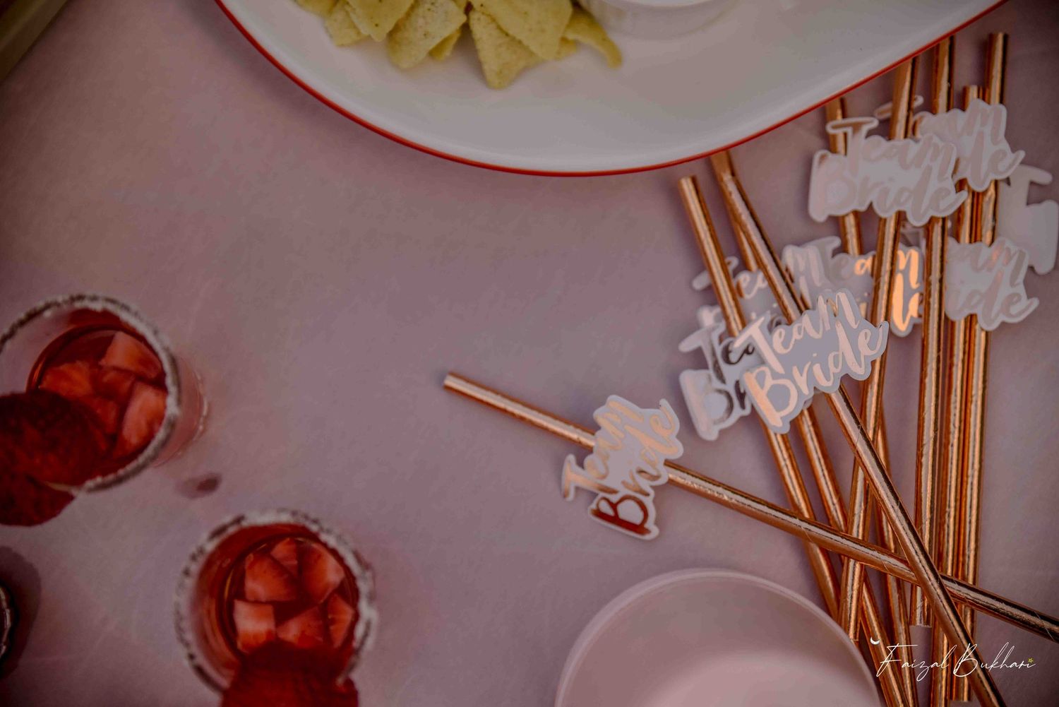 Decorative gold straws with bride-themed toppers beside strawberry drinks on pink tablecloth.