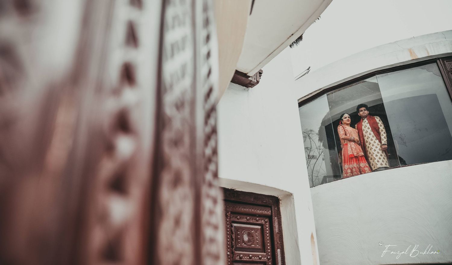 Creative architectural shot of Indian wedding couple in pink attire reflected in curved window beside carved door.