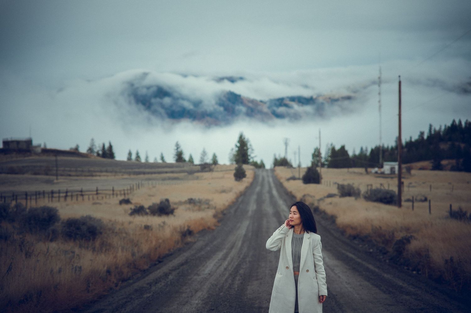 A woman in a white coat stands on a rural dirt road with dramatic cloudy mountains in the background. Clicked by top portrait photographer