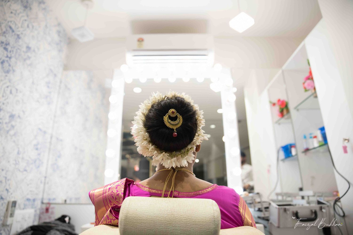 Indian bride sits in salon chair with traditional floral bun hairstyle adorned with jasmine flowers and gold accessories.
