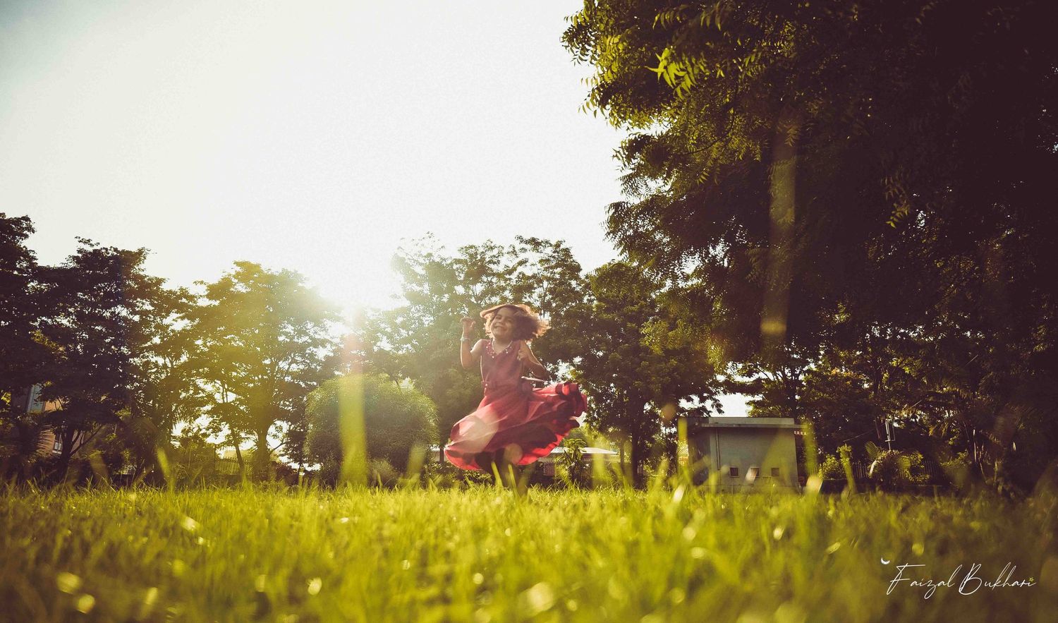 A child in a red dress plays in a sunlit green park with warm golden lens flare in the background.