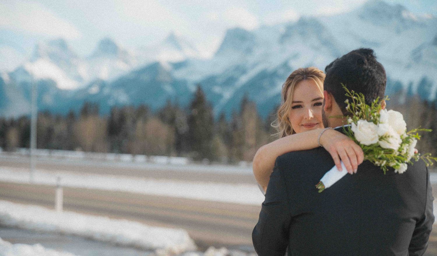 Bride in white off-shoulder gown hugging groom with white rose bouquet against snowy mountain backdrop.