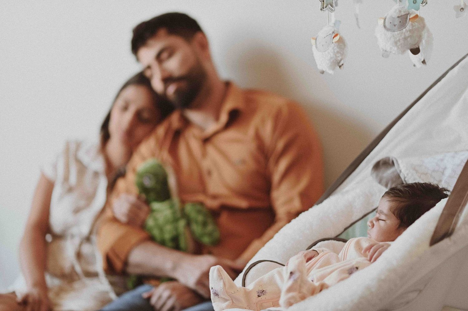 Couple sits together holding a green stuffed toy while newborn sleeps in swing nearby.