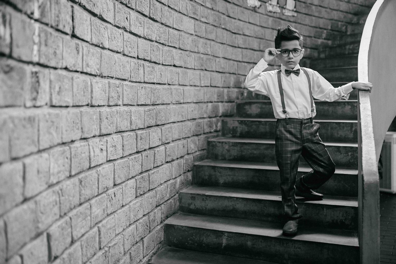 Fashionable boy in glasses, bow tie and suspenders posing on brick staircase in black and white.