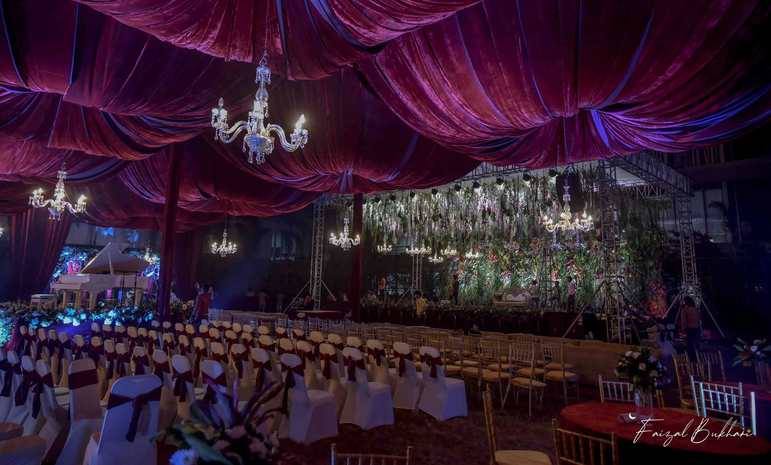 Luxurious Indian wedding hall with deep purple velvet draping, crystal chandeliers and lush floral stage decor.