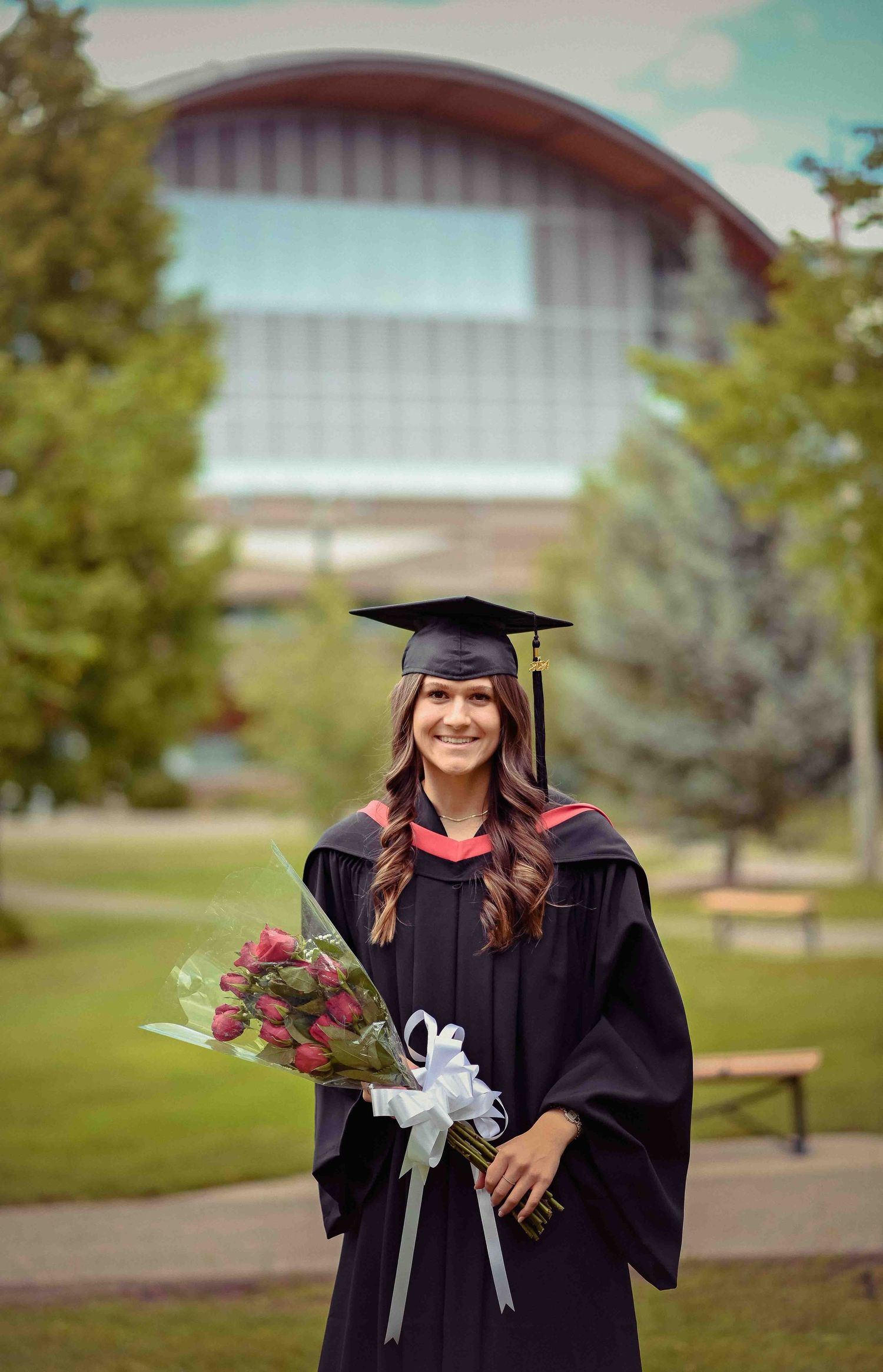 Smiling female graduate in black cap and gown holding red tulip bouquet on college campus green lawn.