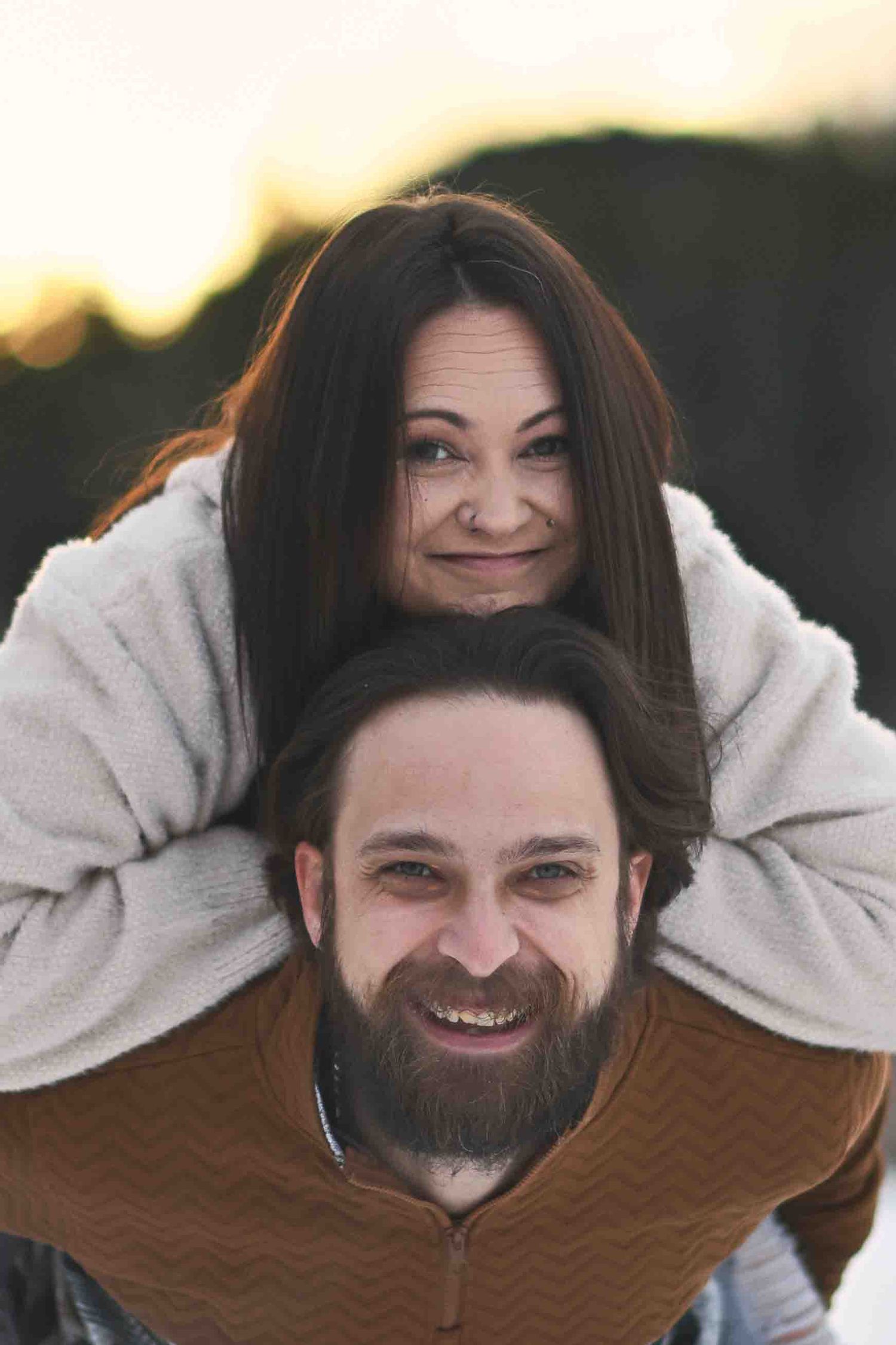 Smiling couple poses playfully outdoors at sunset, woman resting chin on man's head in piggyback.