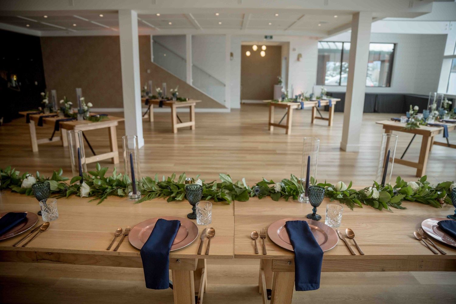 Elegant wedding reception hall with long wooden tables, navy blue napkins, greenery garlands and candlestick centerpieces.