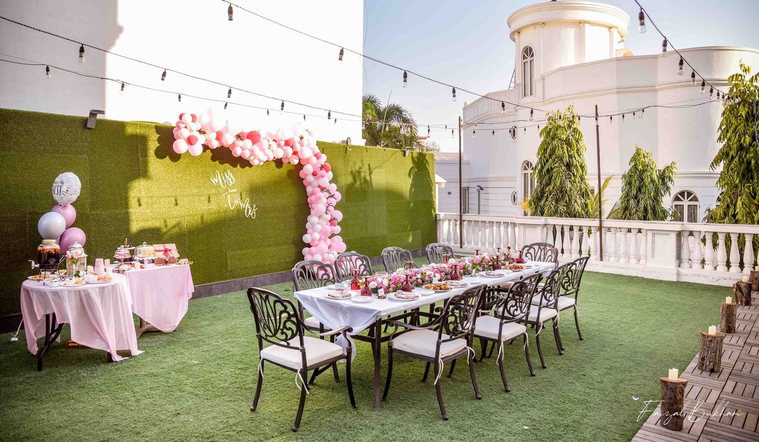 Rooftop bridal shower venue with long dining table, pink balloon arch, and string lights against green wall.