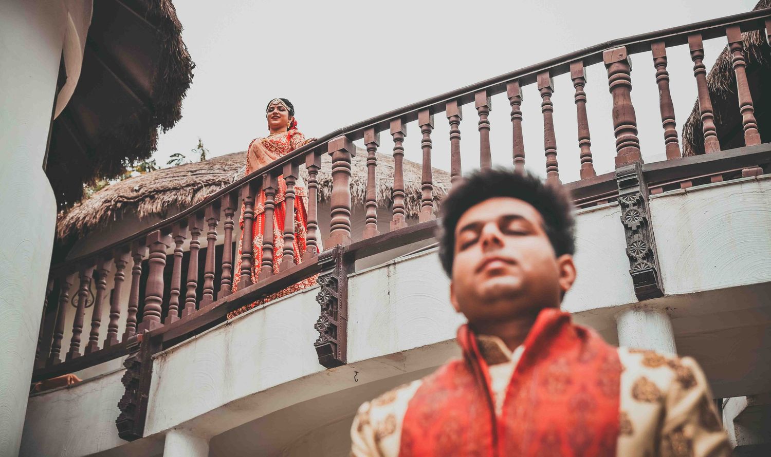 Indian wedding couple captured on ornate wooden staircase balcony in traditional red and gold ethnic outfits.