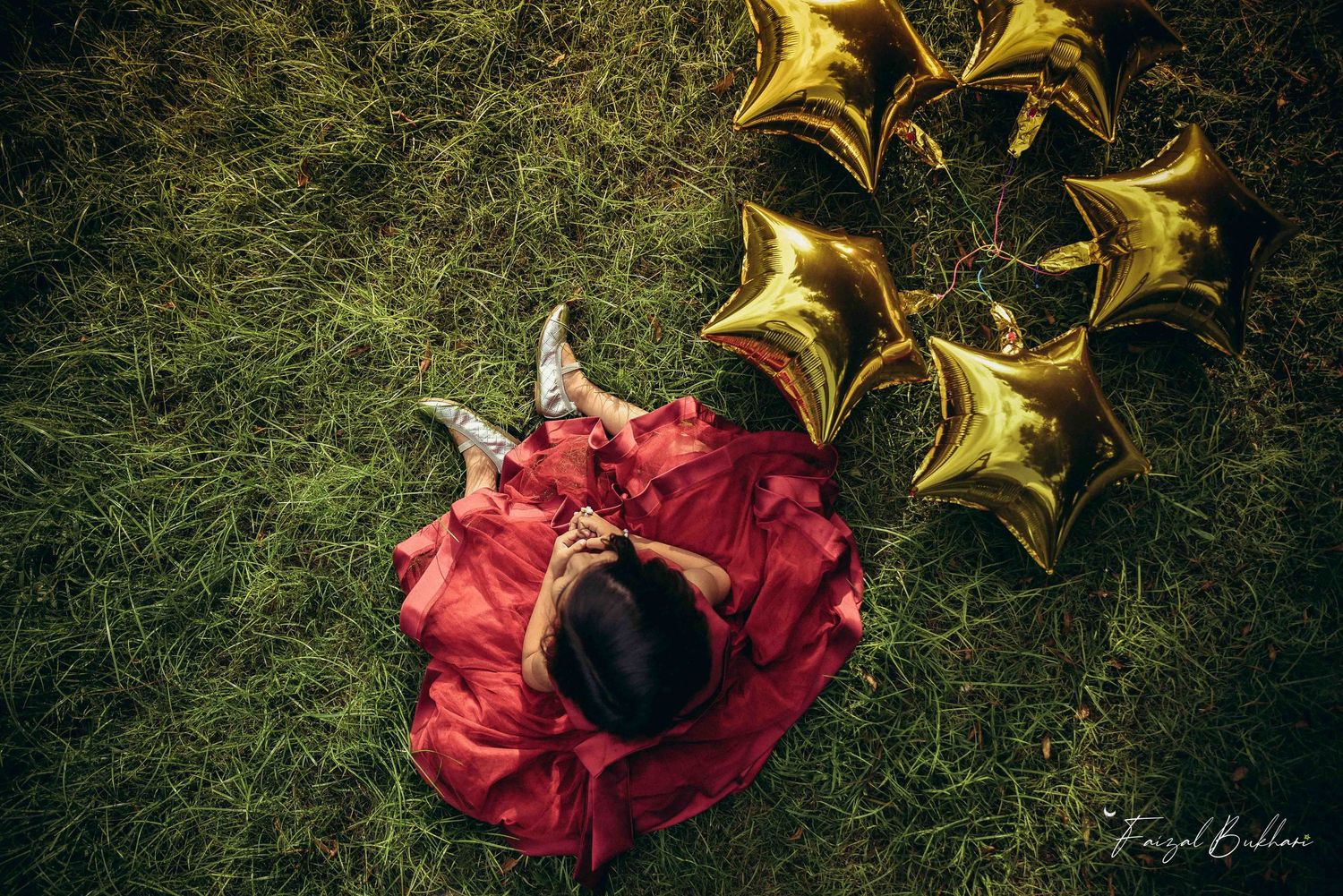 An overhead view of a young girl in a red dress lying on grass beside gold star-shaped balloons.