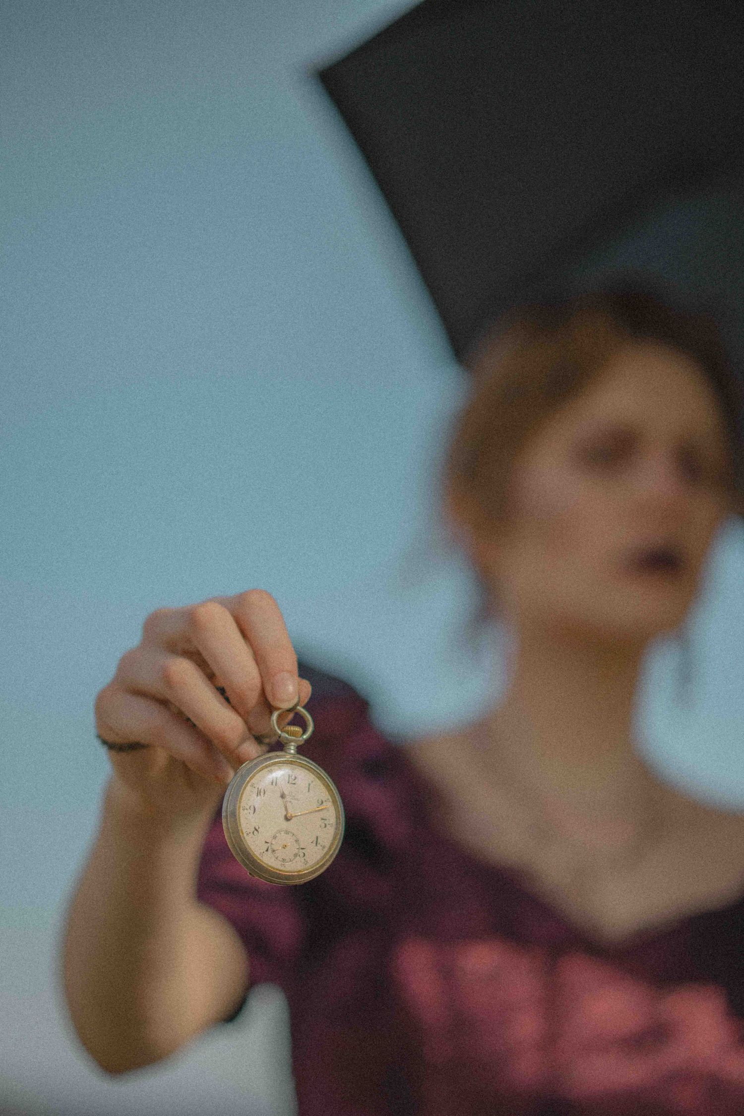 Woman in burgundy dress holds vintage pocket watch toward camera while holding black umbrella outdoors.