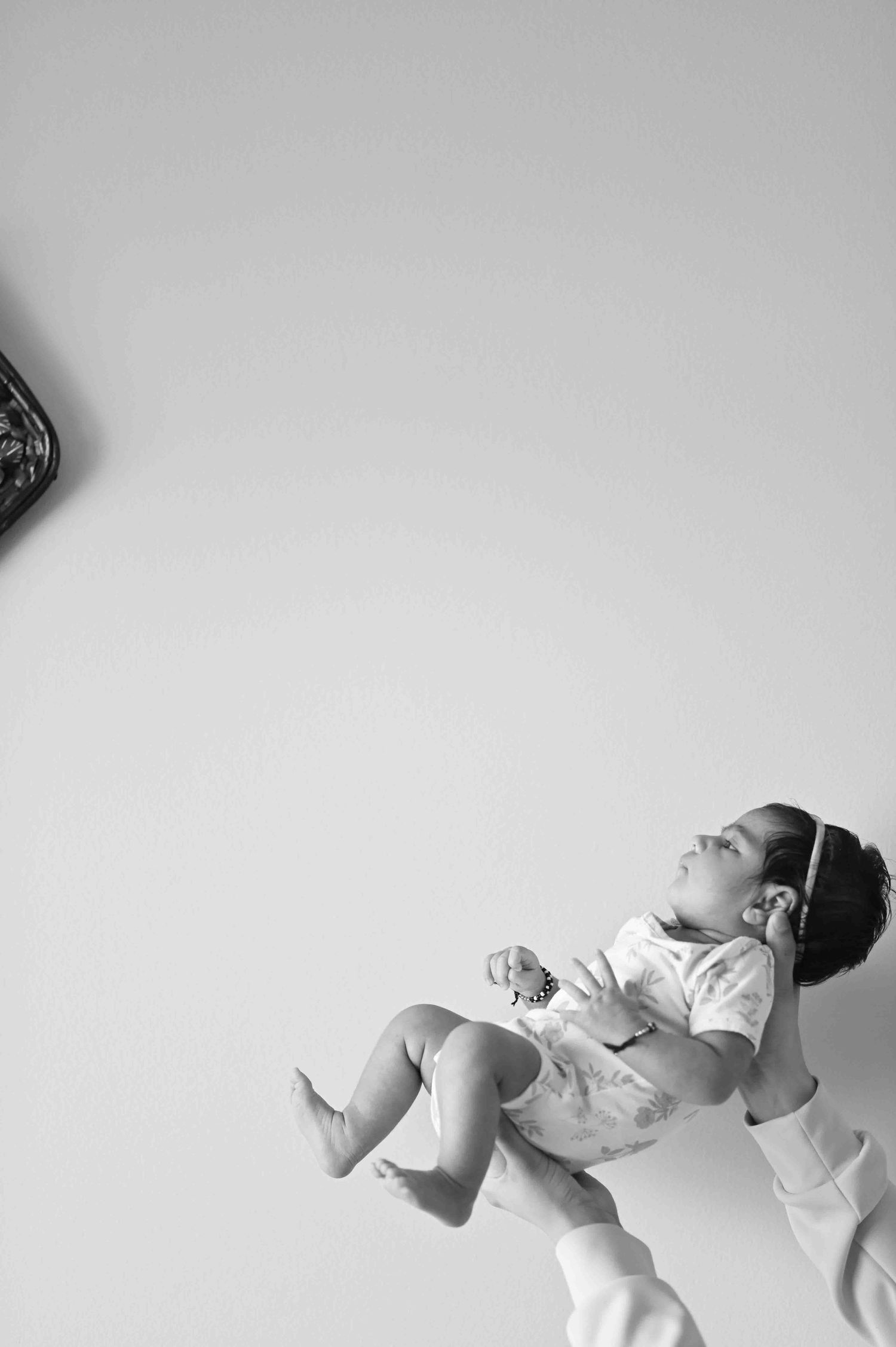 Black and white photo of baby being held up by parent against plain white background, looking upward.
