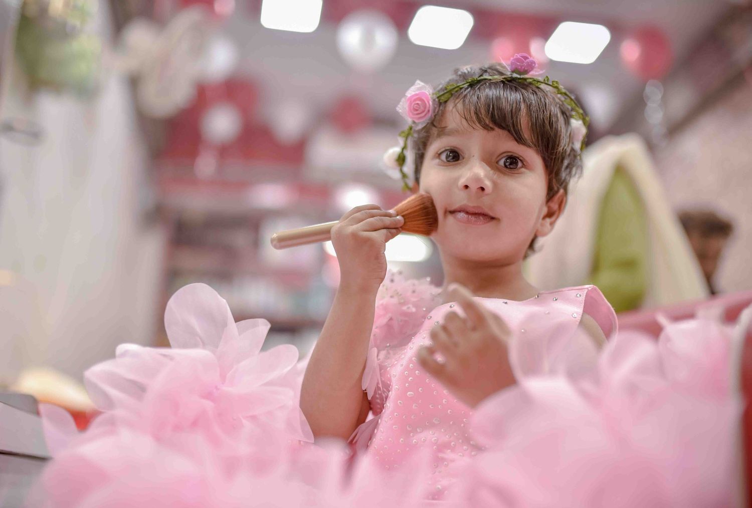 Young girl in pink tulle dress applying makeup brush while sitting at a vanity station.