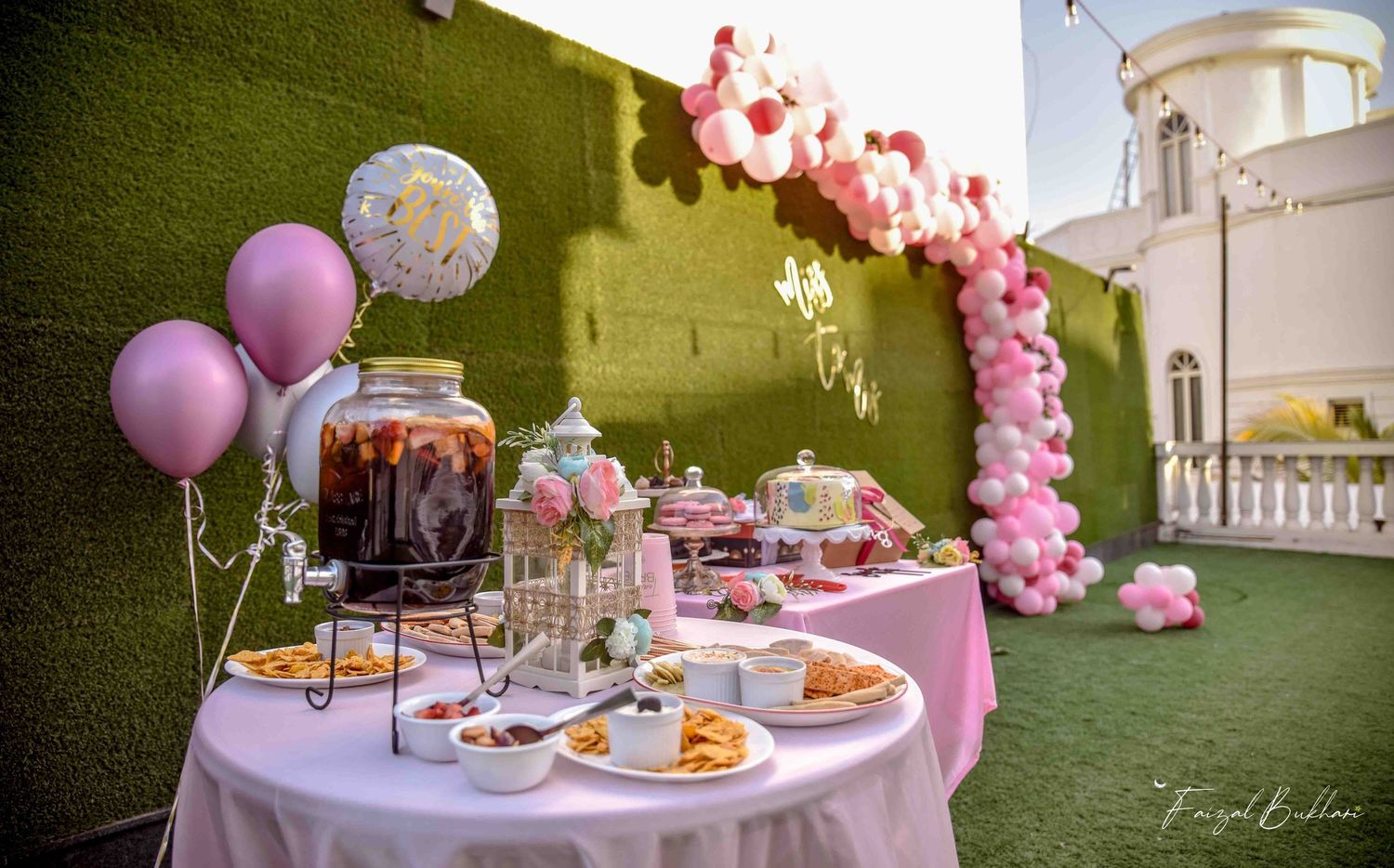 Pink-themed bridal shower dessert table with balloon arch, beverage dispenser, and assorted treats outdoors.