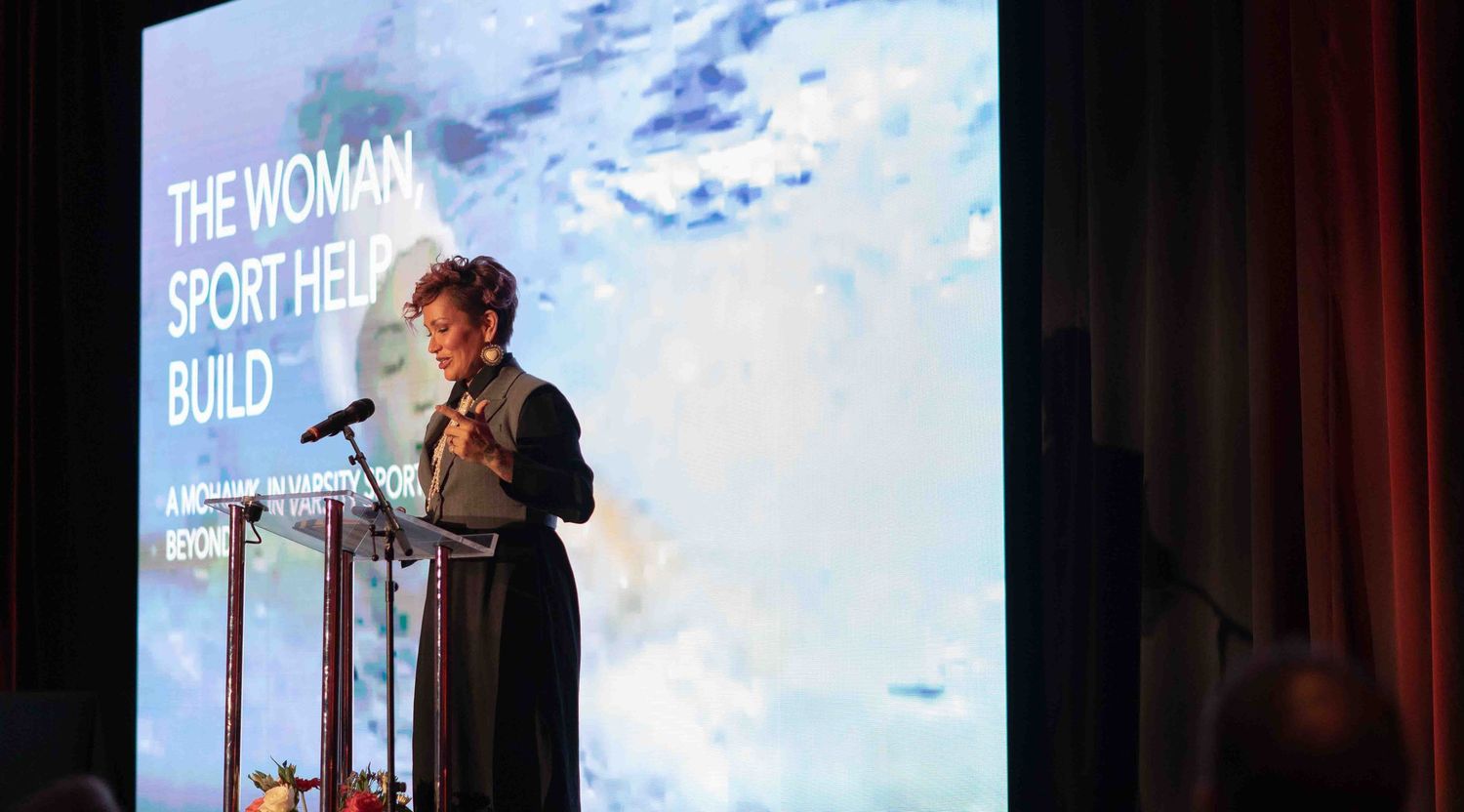 A speaker presents at a podium beside a screen reading The Woman Sport Helped Build at a formal event.