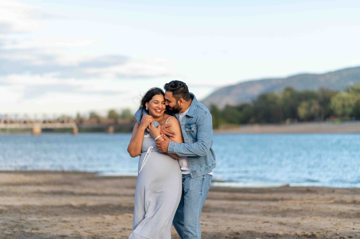 Couple embracing on sandy beach shore with lake and mountains in background.