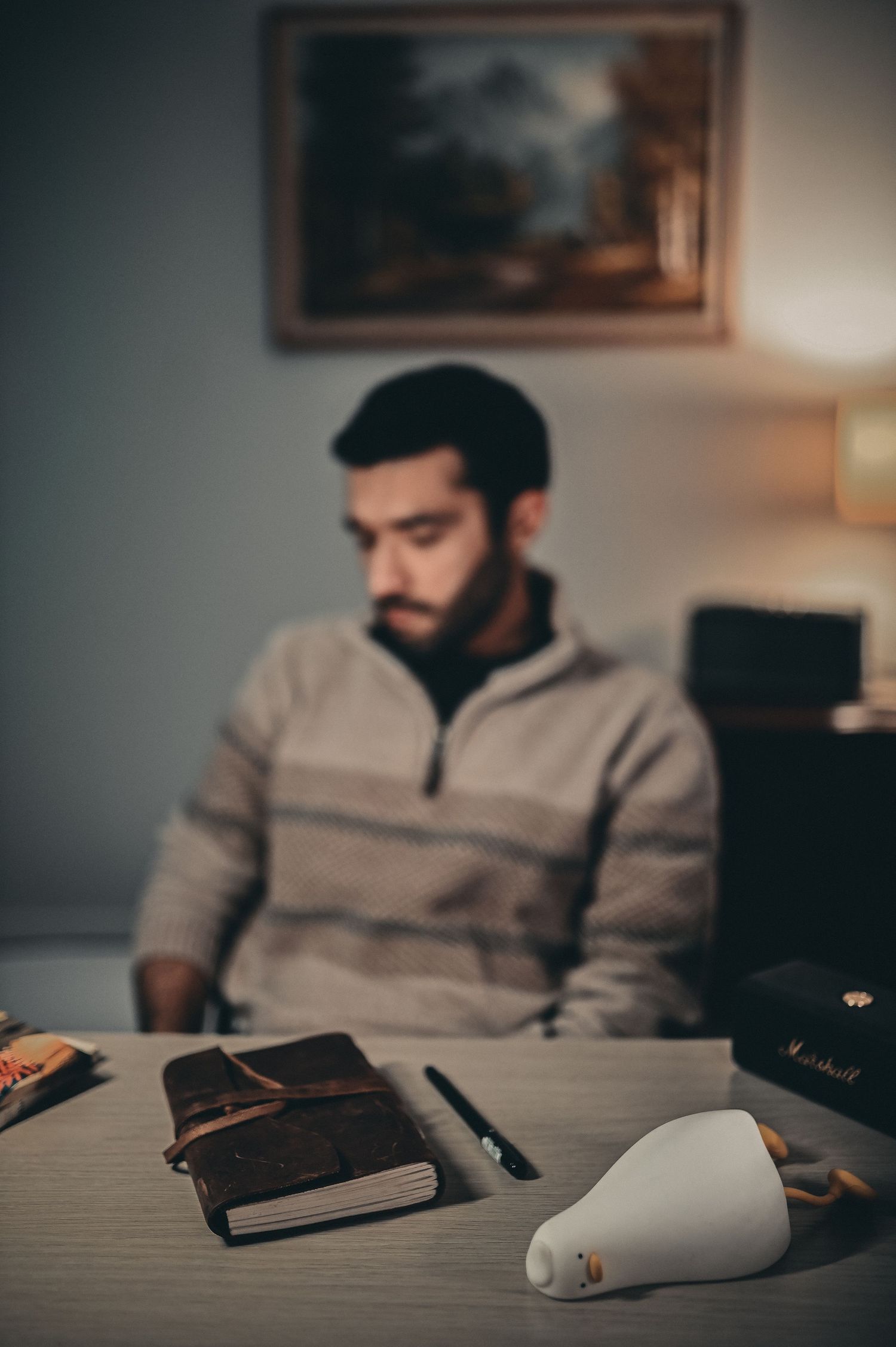 Bearded man in grey sweater sitting at desk with leather journal and lamp in dimly lit room.