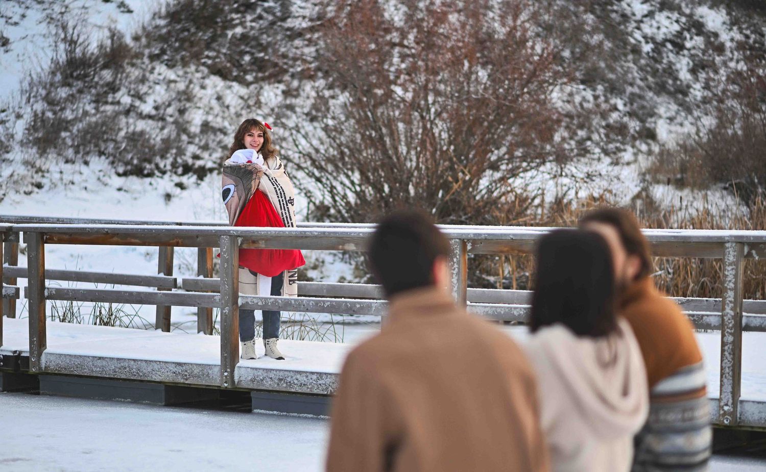 Woman in red skirt poses on snowy bridge while photographers capture her winter portrait outdoors.