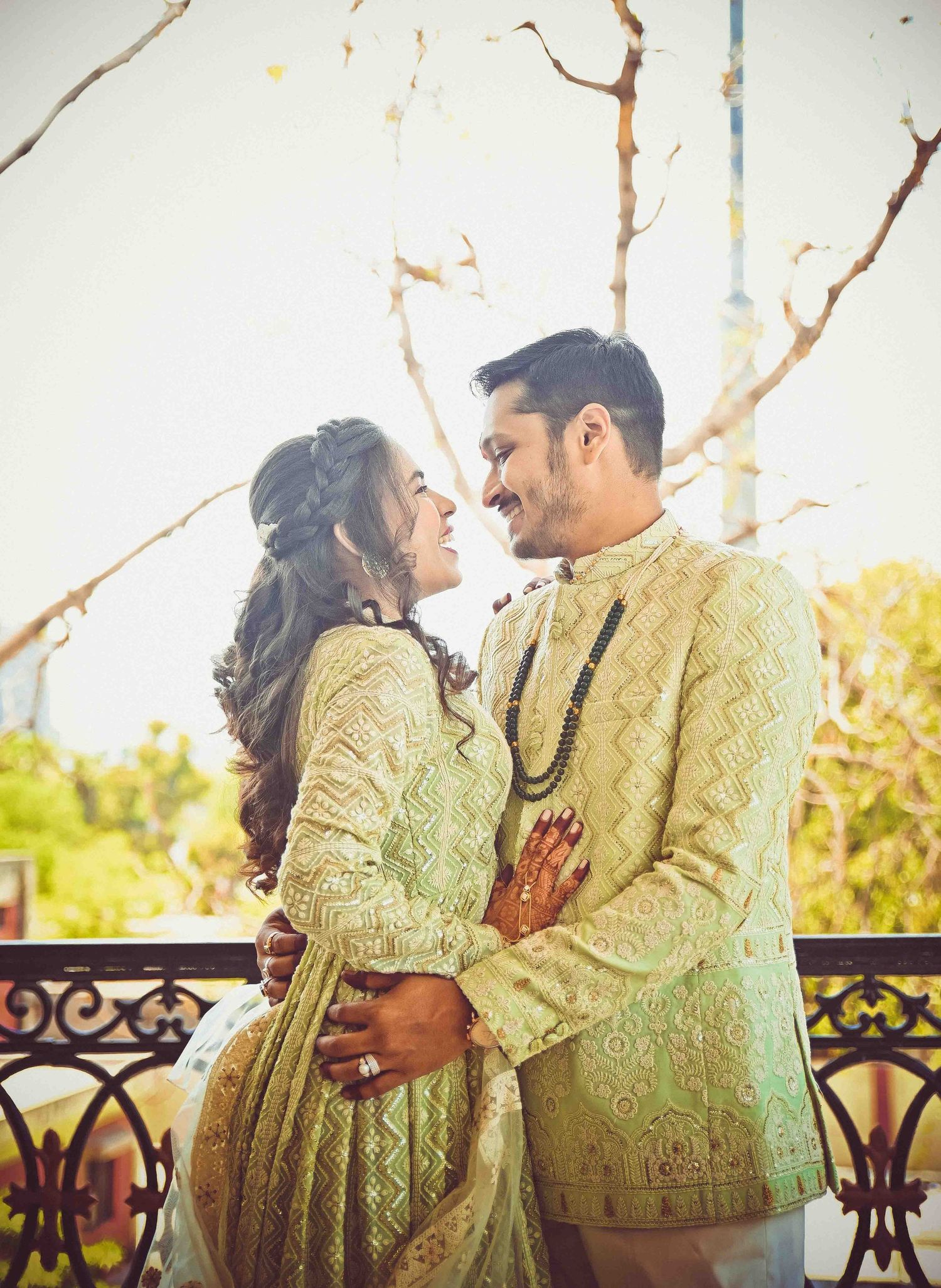 A smiling Indian couple in matching mint green outfits share a romantic moment on a decorative balcony.