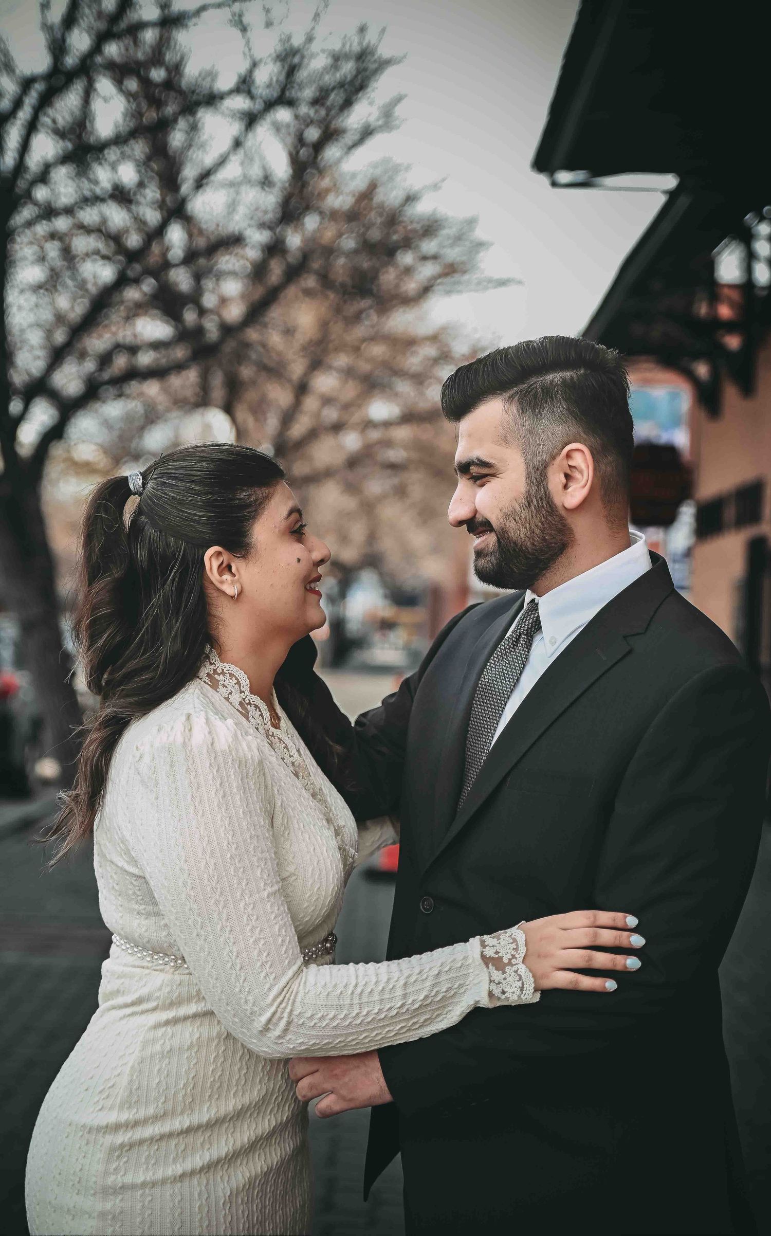 Couple shares a romantic moment outdoors in winter, man in suit and woman in white lace dress smiling.