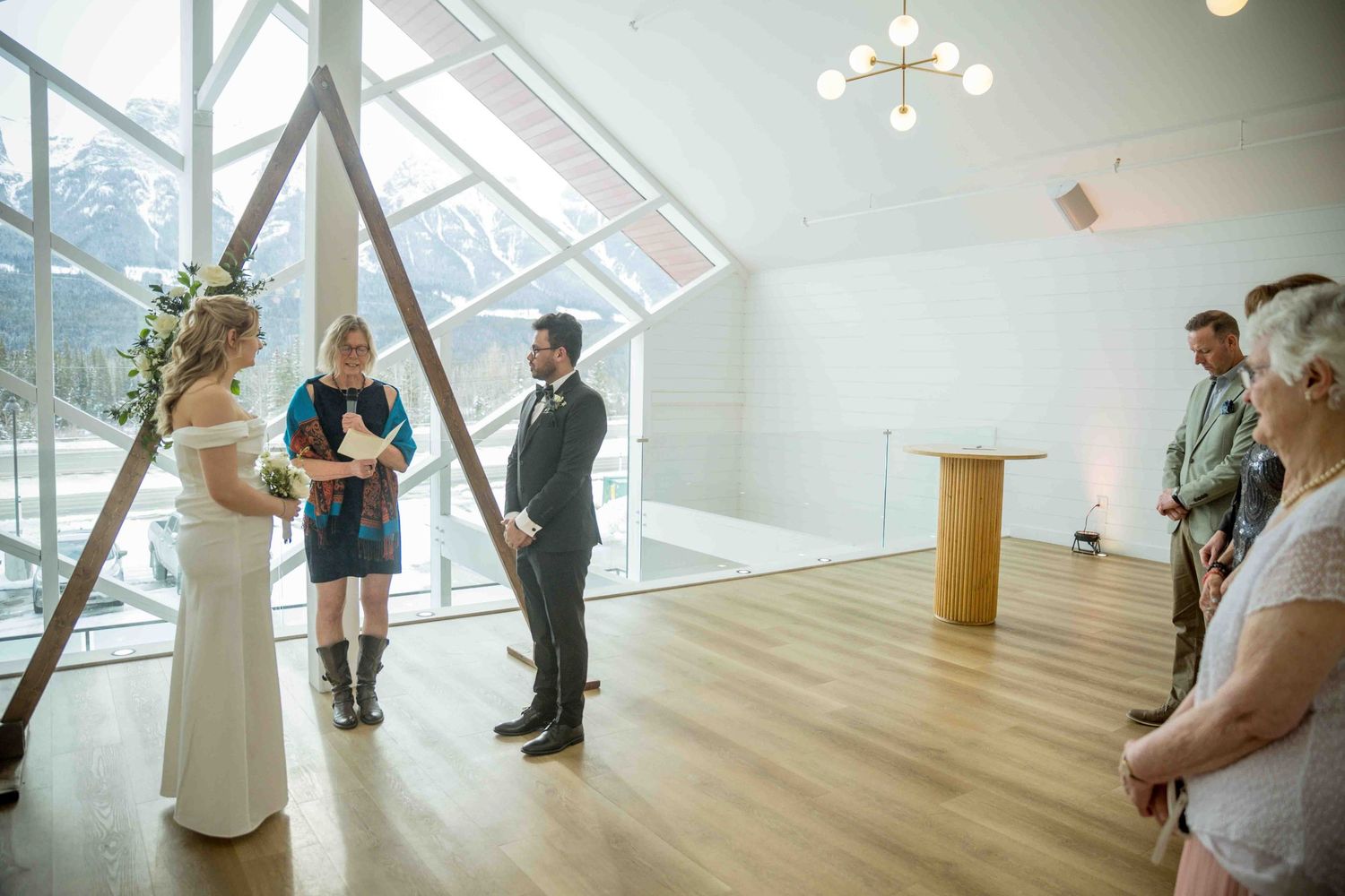 Wedding ceremony inside modern venue with A-frame arch, mountain backdrop, bride in white gown and groom in gray suit.