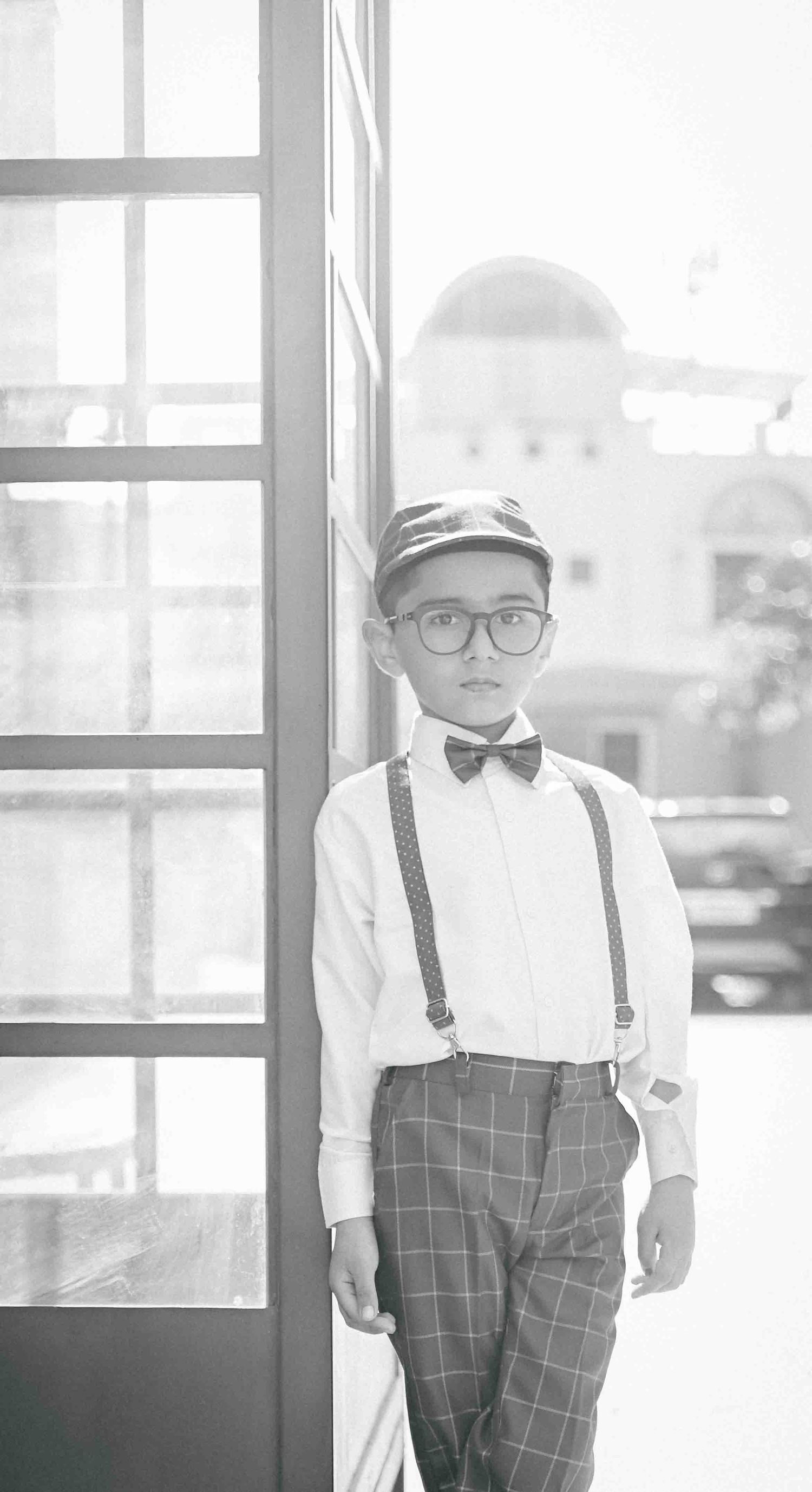 A neatly dressed boy in suspenders, bow tie, plaid pants, and cap stands by a glass door outdoors.