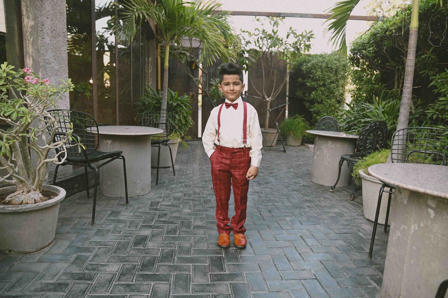 Dapper young boy in white shirt, red suspenders and bow tie walking on cobblestone patio.