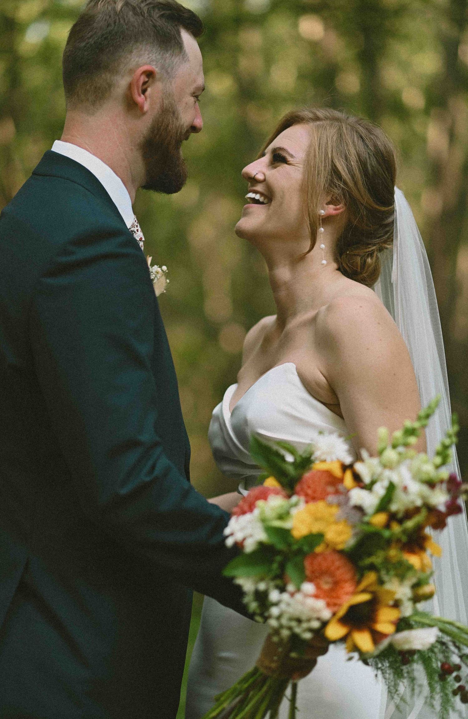 Bride and groom laugh together in forest, bride holding vibrant sunflower and wildflower bouquet in wedding attire.