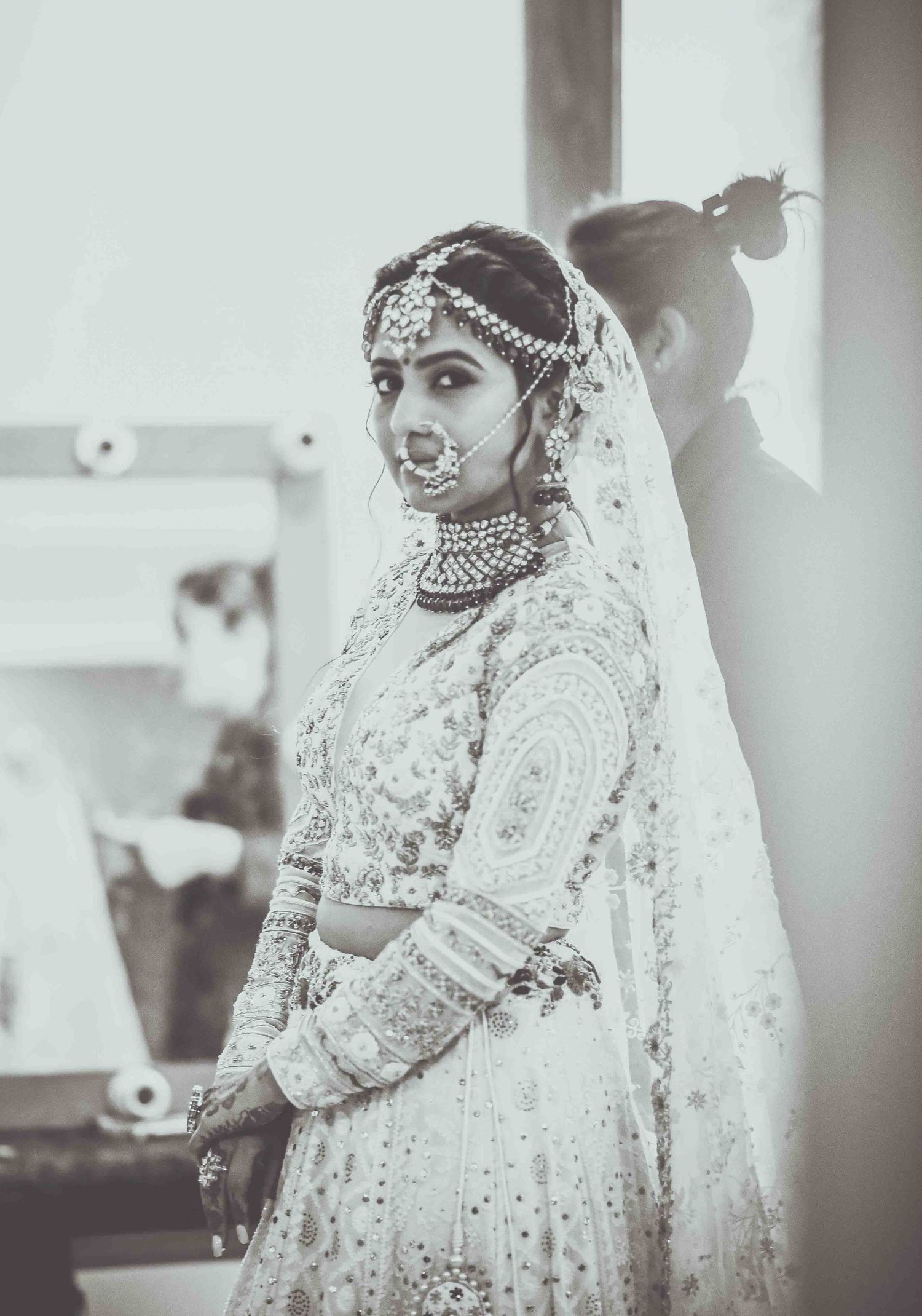 Elegant Indian bride in white embroidered lehenga with maang tikka, nose ring and veil in black and white photo.