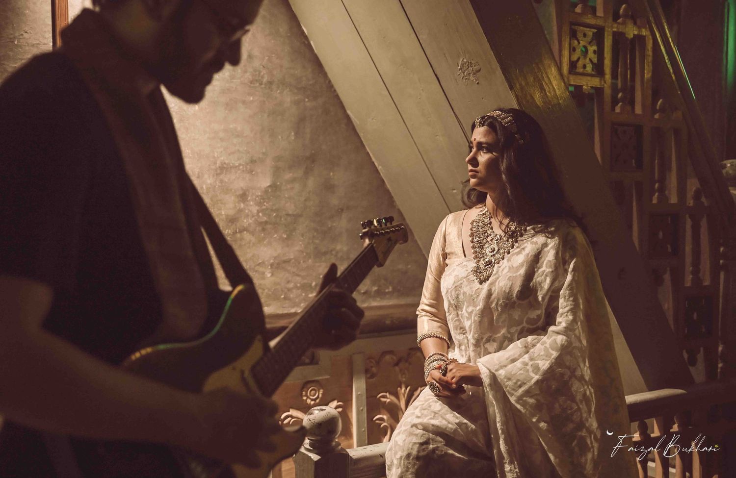 Bride in white embroidered lehenga listening to guitarist in a warmly lit artistic indoor setting.