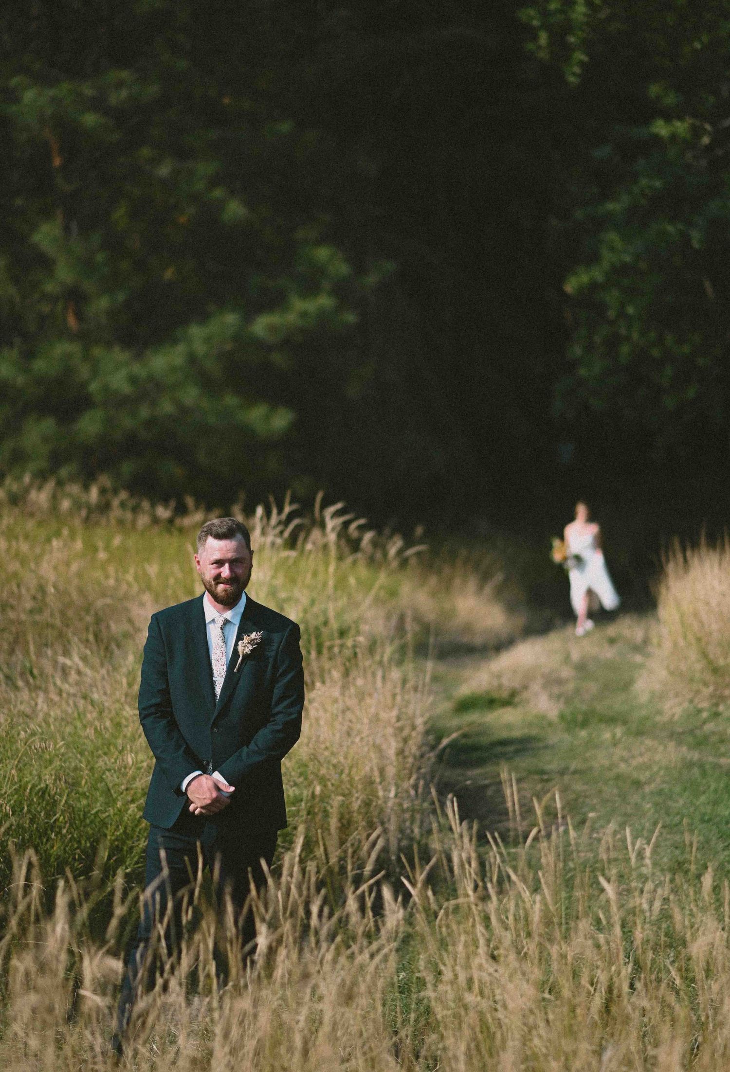 Groom in dark suit stands in golden meadow as bride in white dress approaches along a forest path.