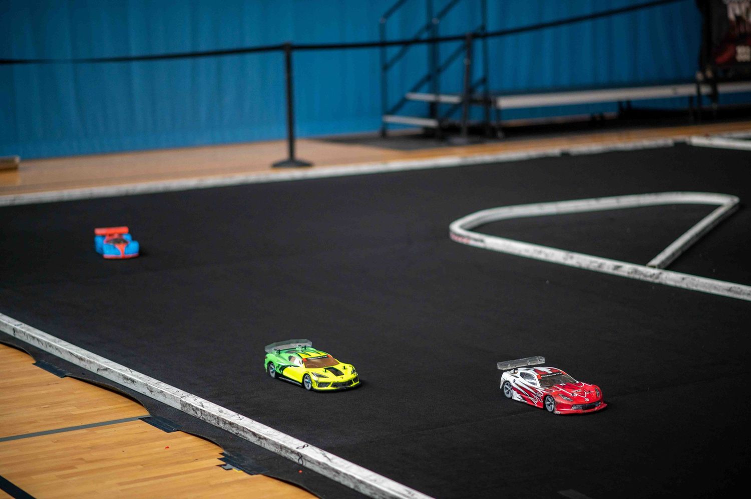 Green and red RC cars racing on a large indoor black carpet track.