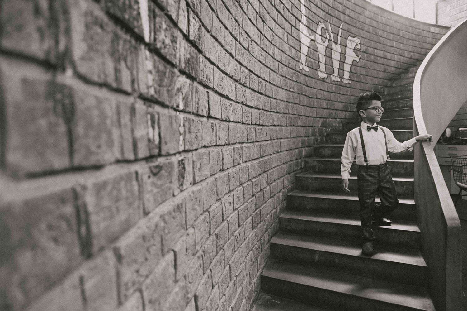 Small boy in suspenders descending brick staircase beside curved graffiti wall in black and white.