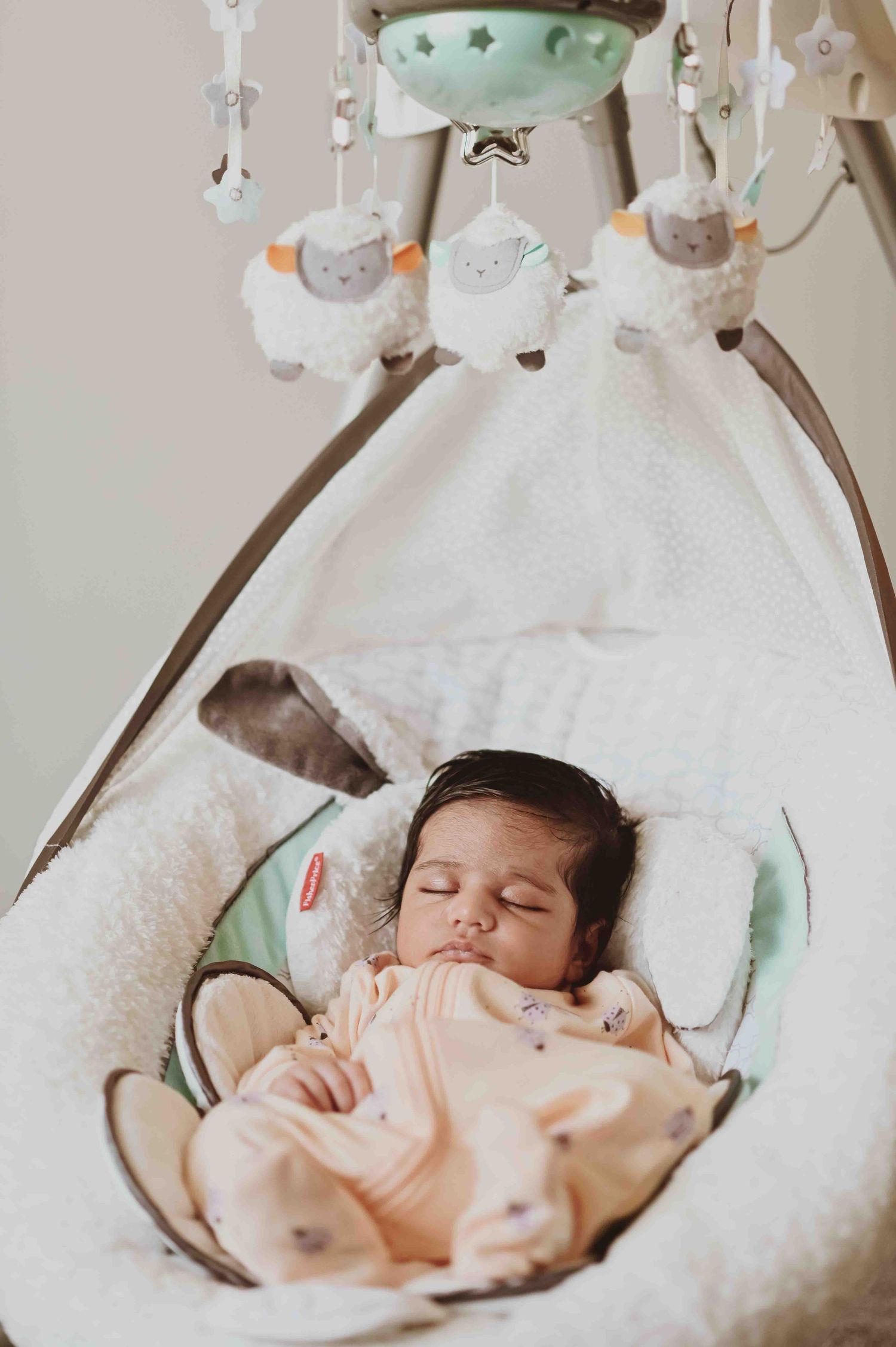 Sleeping newborn baby in a peach outfit rests in a white fluffy baby swing with mobile.