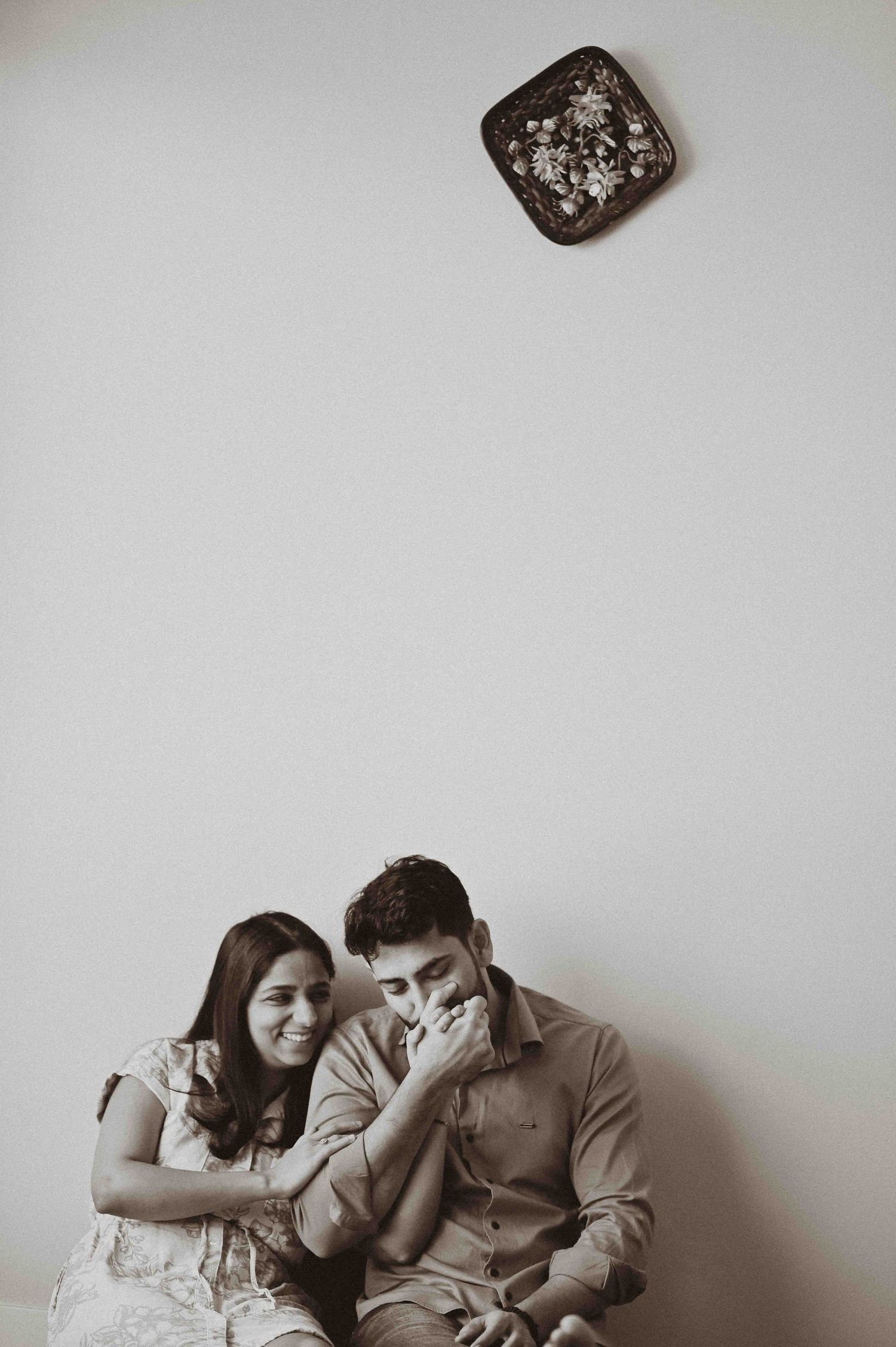 Couple sitting together against white wall in black and white photo, man kissing woman's hand while she smiles warmly.