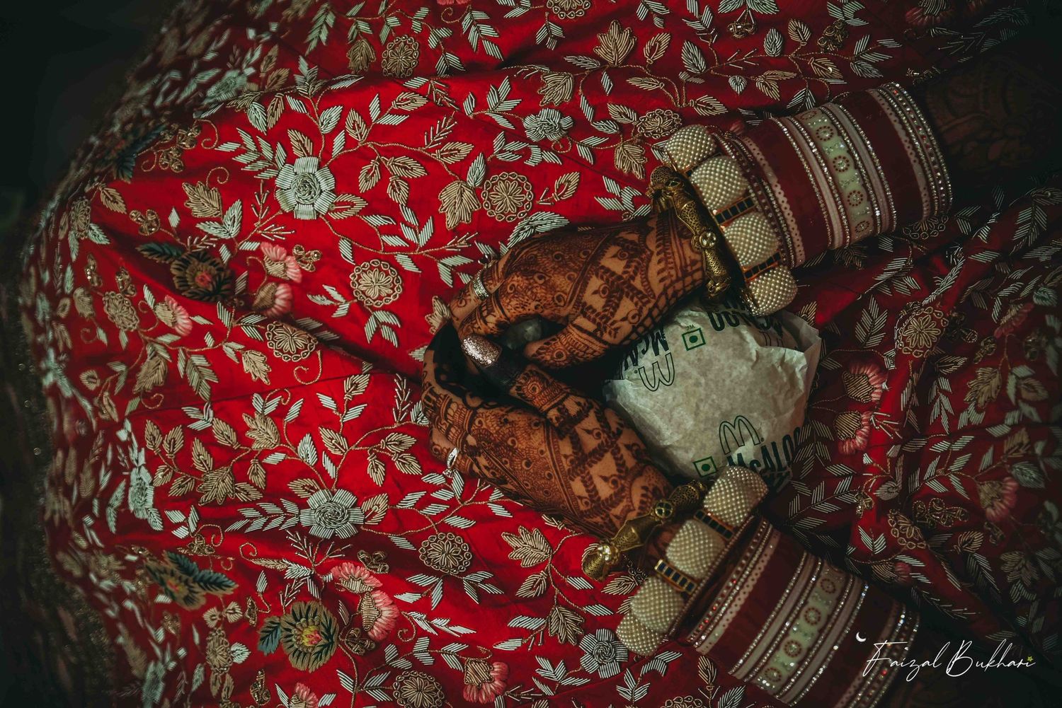 Close-up of bridal hands adorned with intricate mehndi henna designs and gold bangles on red floral lehenga.