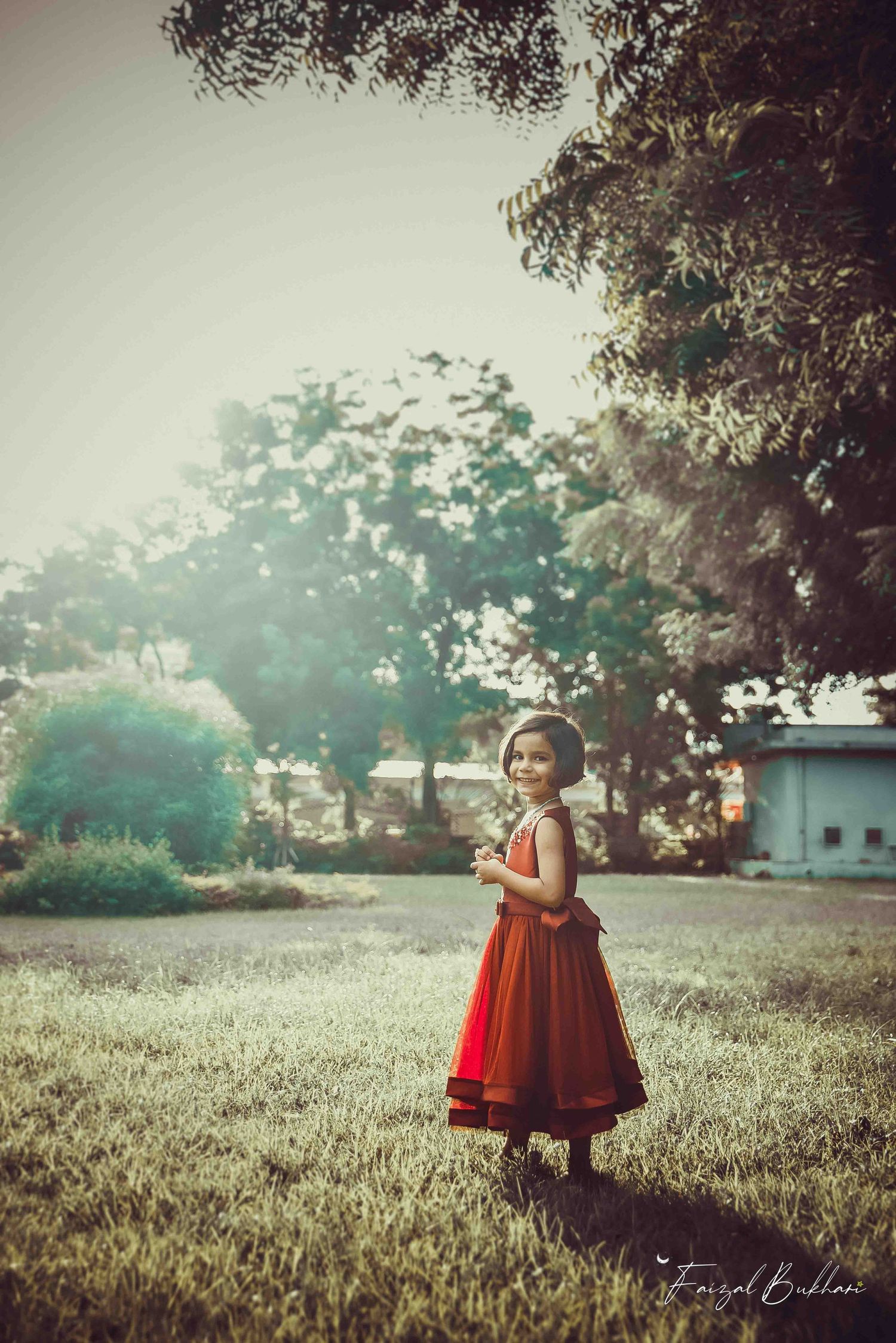 A smiling little girl in a beautiful red traditional dress standing in a sunlit green park.