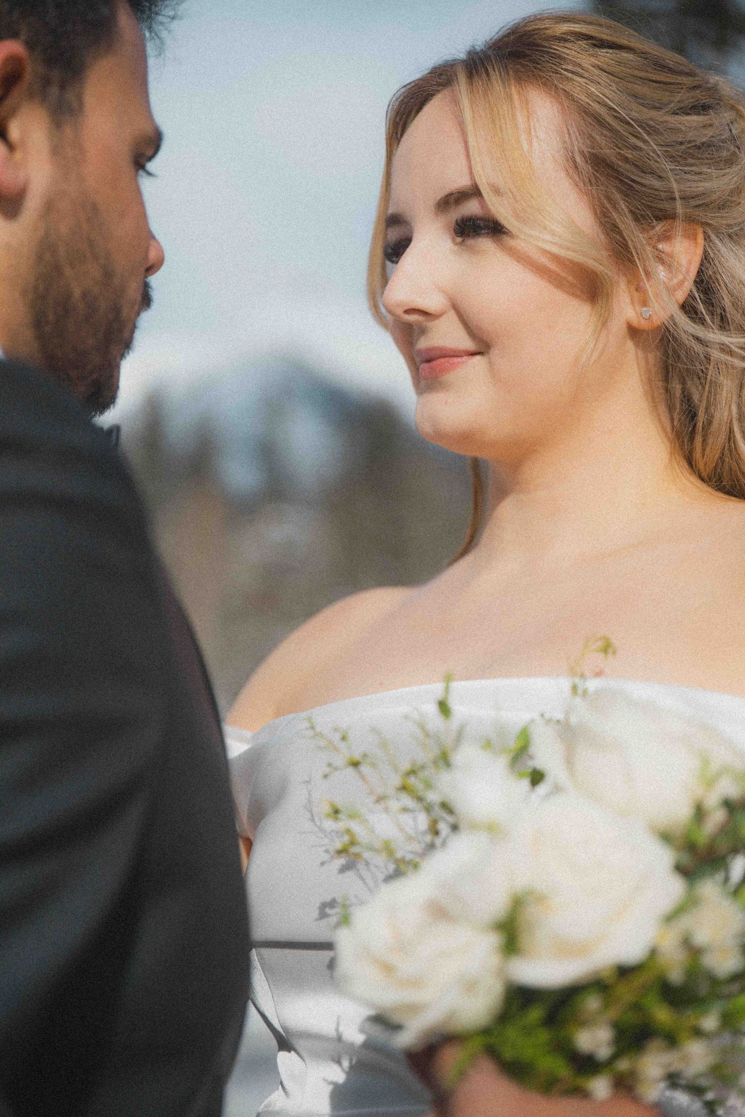 Smiling bride in off-shoulder gown holding white rose bouquet gazing at groom during outdoor wedding portrait.