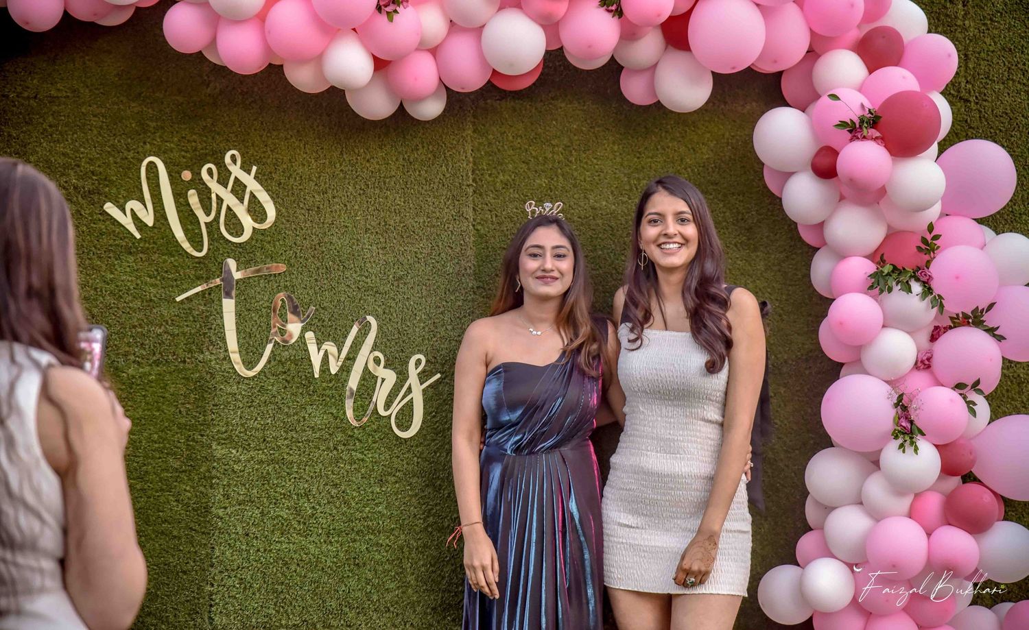 Bride-to-be wearing tiara poses with friend at Miss to Mrs green wall balloon arch backdrop.