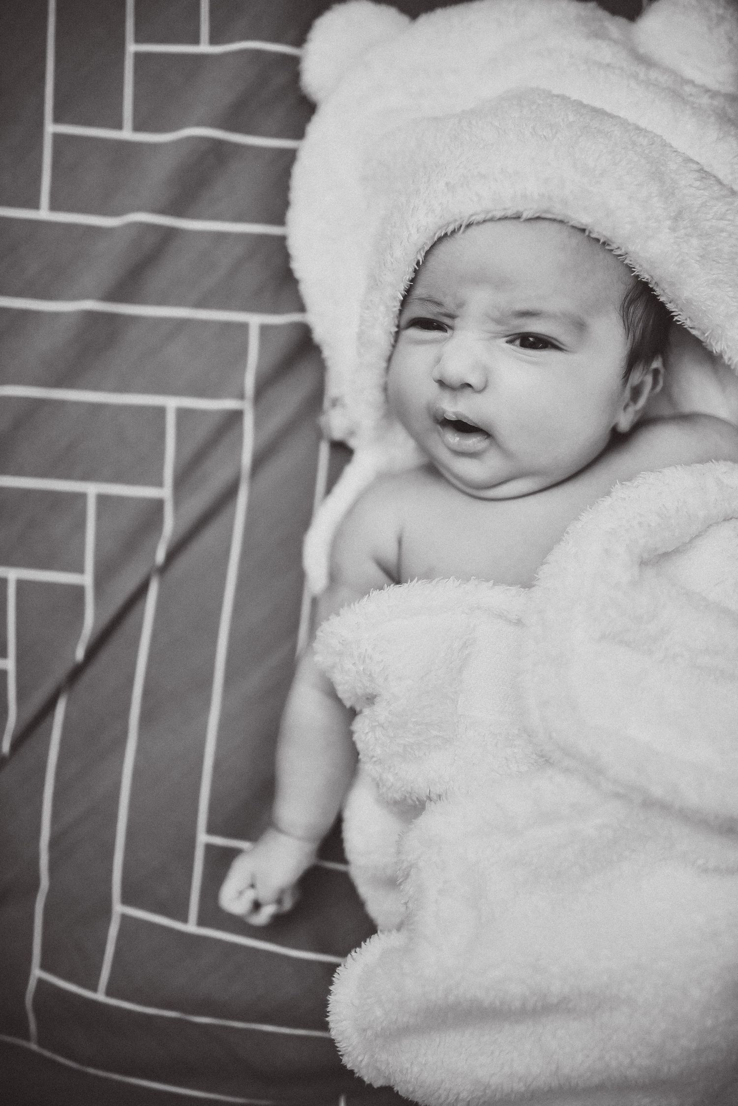 A black and white portrait of a newborn baby wrapped in a fluffy hooded towel lying on a blanket.